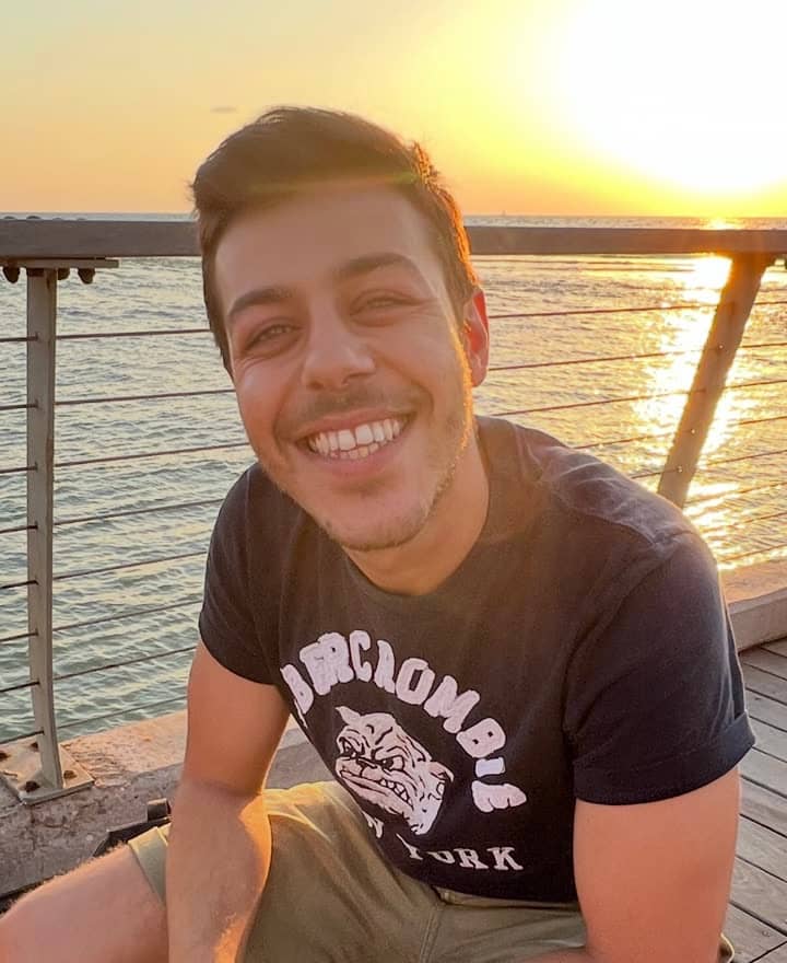 Mert smiling on a wooden boardwalk by the sea at sunset.